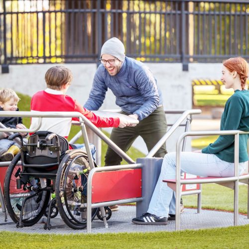 A man stands laughing as he spins the wheelchair-accessible spinner, along with a teenage wheelchair user. Also on the spinner are another teenager and an adult with a young child in her lap, all smiling.