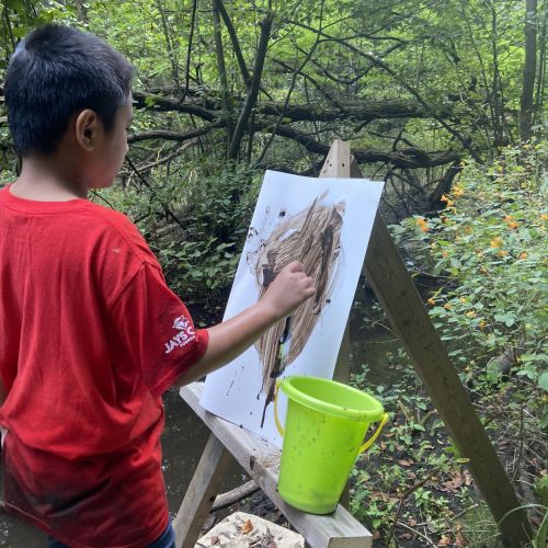 A child in a red shirt is painting outdoors on a white canvas set up on an easel. The scene is surrounded by green trees and foliage. A green bucket is placed on the easel's lower shelf, and there is a stream flowing nearby. The child is painting with browns and other earthy tones.