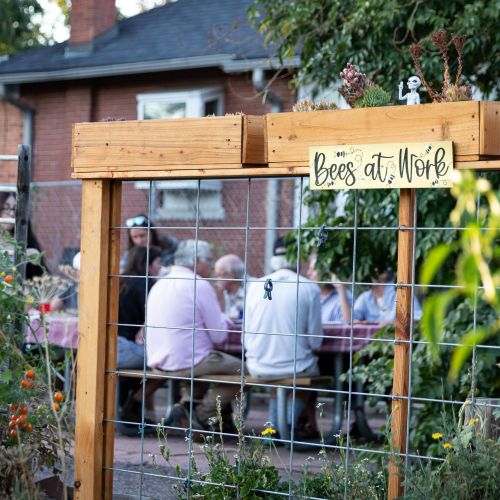 A group of older adult gardeners gather at a table in the background. In front of them is a fence with a sign that says, “Bees at Work”.