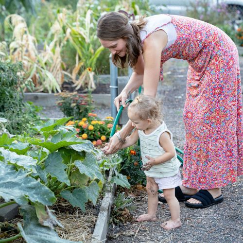 An adult wearing a red and pink patterned dress helps a young child water a row of colorful flowers and vegetables inside the garden.