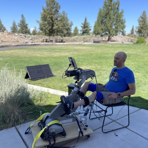 On a patio, a man rides the RT300 FES cycle next to an open green space with trees and shrubbery in the background. Placed in bright sunshine, a solar panel powers the RT300 FES cycle. 