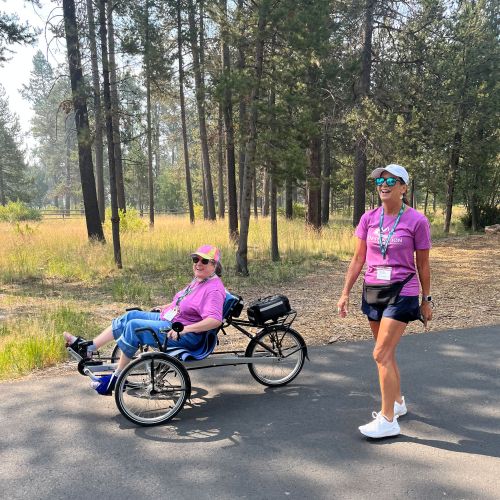 An Adventure Group participant rides a mixed recumbent/upright bicycle, smiling at the camera. She is accompanied by a smiling woman on foot, in front of wooded parkland and grasses.