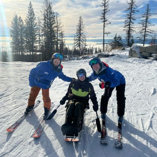 A participant is in a sit ski for a picture, with a DREAM volunteer and a seasonal staff employee posing on either side of her. The scenery behind them is an open ski slope covered with snow that shines bright from the sunlight.