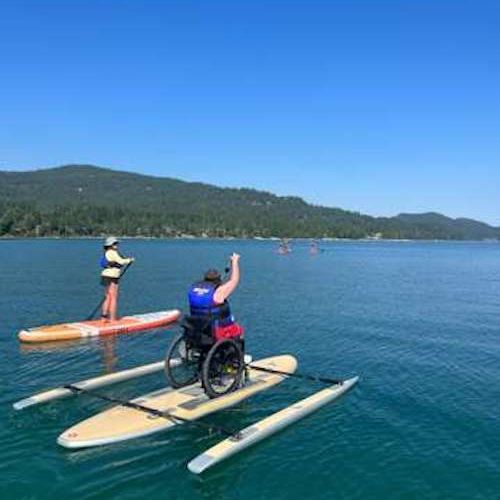 Paddling on Whitefish Lake on one of three OnIt paddleboards in the DREAM watersports fleet. A participant sits in a wheelchair, which is strapped securely on an Onit paddleboard that allows the participant to paddle the board either by themselves or with the help of a volunteer or staff member standing behind them. The outriggers on the Onit make this experience fun and safe and give participants a sense of freedom to allow them to paddle on their own. The outriggers can be removed for an additional challe