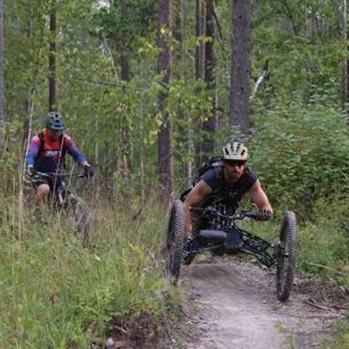 Participant comes down off a mountain bike trail in a handcycle. An eager participant comes down off a mountain bike trail, with a volunteer instructor following close behind on a mountain bike to ensure a smooth, safe, and fun ride. Trees and foliage line the narrow bike trail. The participant himself is riding a hand cycle, which is one of the many adaptive bikes that DREAM provides to accommodate a large variety of disabilities and needs.