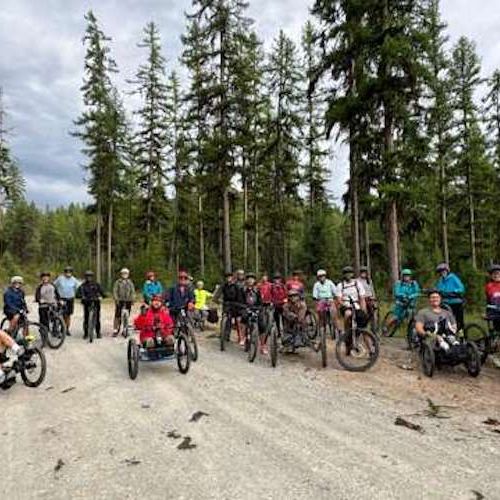 A group photo from one of DREAM's many Summer Tuesday mountain bike rides. Everyone enjoys a breezy, cloudy day as they ride along a bike path bordered by pine trees. In our inclusive group model, participants ride at their own comfort level and feel challenged to try new skills on their adaptive three-wheeled bikes. Riders are accompanied by veteran participants, volunteers, and staff.