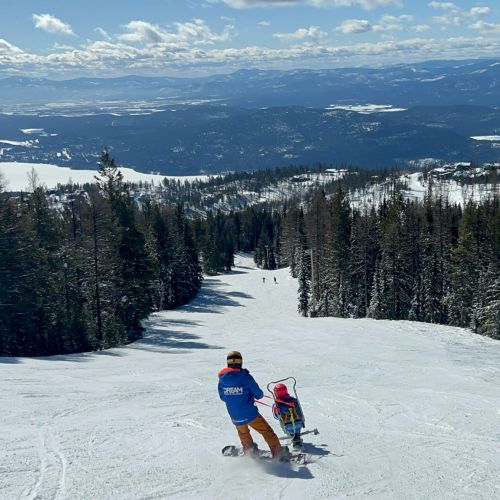 The view from the top of Whitefish Mountain Resort, where a ski instructor and participant stand before a snow-covered ski slope that is bordered by dark pine trees. Clouds line the blue sky above them. The participant's sit ski is tethered to the instructor, which helps them learn and hone ski skills such as lean and turn while staying close to the instructor.