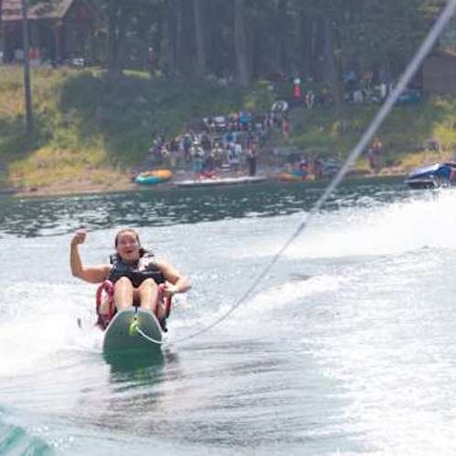 Sit water ski on Echo Lake- A participant water-skis across a lake, shouting gleefully and raising a triumphant fist in the air. They are using a sit ski, which can be used with or without outriggers as is shown in this picture. This adaptability provided by DREAM allows participants to enjoy water skiing at their own comfort level and also allows for skill building of the sit water ski sport.