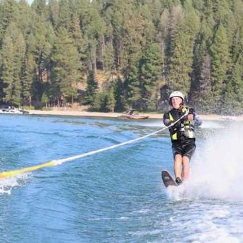 The participant waterskiing on a tree lined lake has a visual and hearing impairment. He is equipped with a waterproof headset that allows him to receive instruction from the boat staff while he is waterskiing.