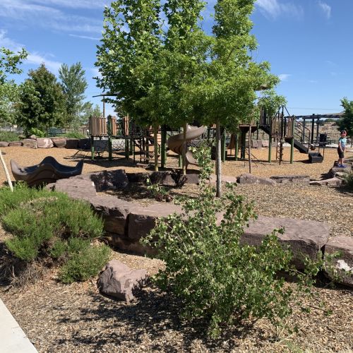 A concrete path leads to a playground surrounded by plants. An adult who is sitting and young child who is standing are in the background.