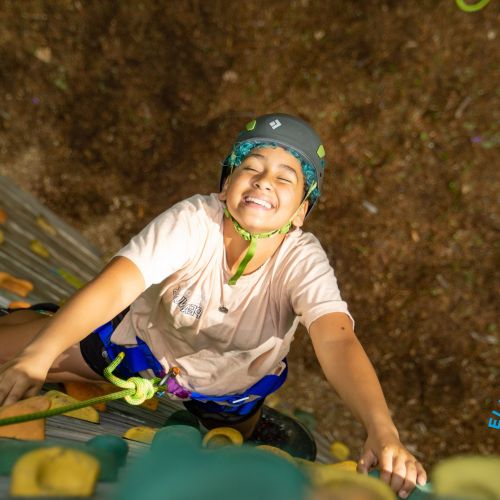 An overhead photo of an Elevate Youth member outdoor rock climbing at Hammond Pond looking toward the top of the route and smiling.