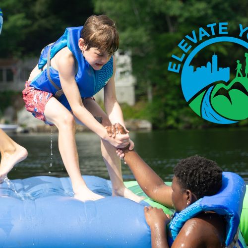 Elevate Youth having fun in the lake during summer camp. One youth is giving another youth a hand onto a blue and green inflatable raft on the lake. 