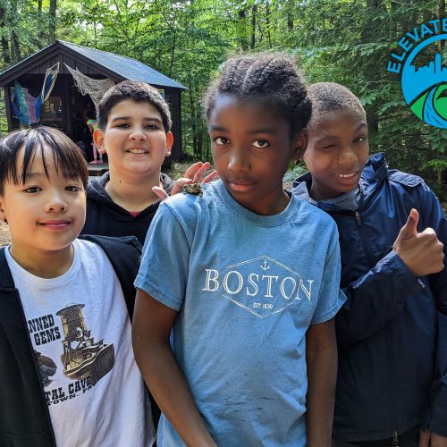 Group of kids standing in the woods at Elevate Youth Summer camp. One youth is winking and giving a thumbs up sign while three other kids smile. A wooden camping structure is in the background amid a green forest.