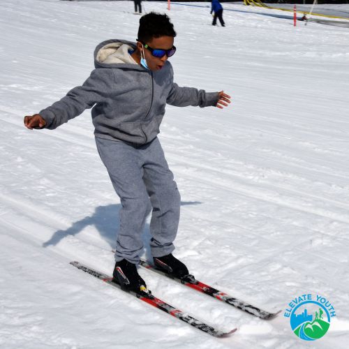 Elevate Youth participant cross country skiing. A child wearing sunglasses stares down at their cross country skis in concentration while skiing on a smooth open stretch of snow on a bright day.