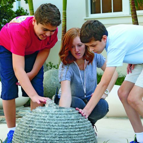 A teacher and two young people crouch down to touch a stone water sphere, with a small fountain of water on the top and ribbed sides, providing both proprioceptive and vestibular sensory experiences. In the background, there are also smooth rocks that offer varied tactile engagement.