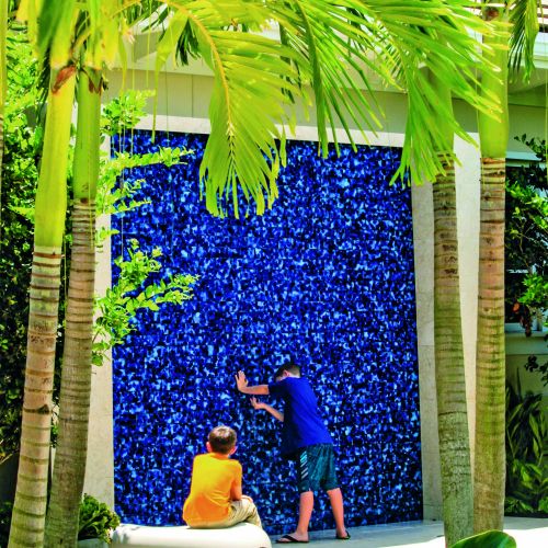 Viewed through the palm trees, a child stands with both of their hands pressed into the water wall, a large blue feature wall offering sensory opportunities, while another looks on from their seat on a nearby rock.