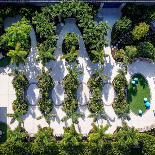 An aerial view of the 13,000-square-foot garden. In an organized pattern, the paths wind around trees and green areas with a building on the left-hand side and a pergola on the opposite end. The log tables and multiple seating areas can also be seen.