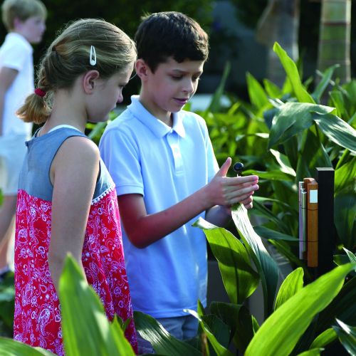 Two children stand at a musical sculpture while one holds the rubber mallet for the instrument. They are surrounded by foliage. In the background, another child can be seen exploring the area.