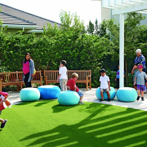 In an open green space, several children play on large blue cushions while adults mill around. Behind this area are two large, curved benches in front of tall green hedging. A pergola provides some shade.