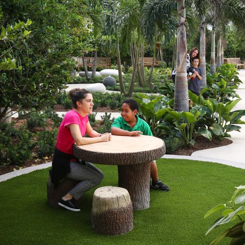 In a nook nestled within lush planting and encircled by a distinctive paving band, a teacher and child sit at a table and stools constructed of rustic logs, smiling and chatting.