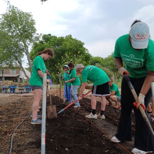 Seed Crew students diligently plow a row of soil in preparation for seed planting at Conundrum Farms. Lined up behind them are other already plowed rows. In the background are tall trees and a farm building; the sky above them is cloudy, which offers plenty of shade.