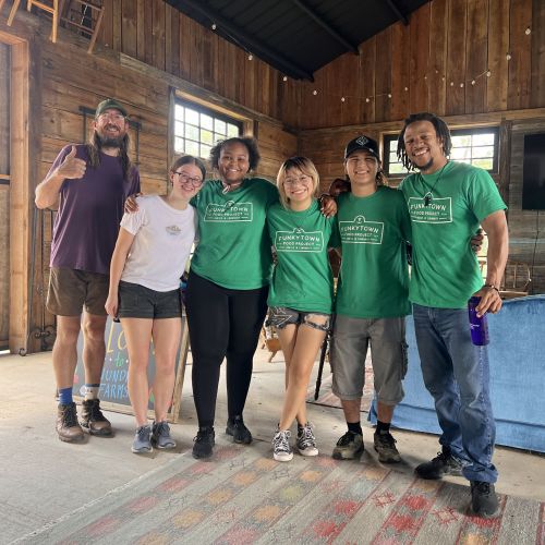  The Program Director Juleon Lewis and Farm Manager Montana Stoval line up for a picture with four Assistant Crew Leaders (ACLs) in a cabin living room. Lewis poses with a thumbs-up and a bright smile, which Stoval and the ACLs also wear on their faces.