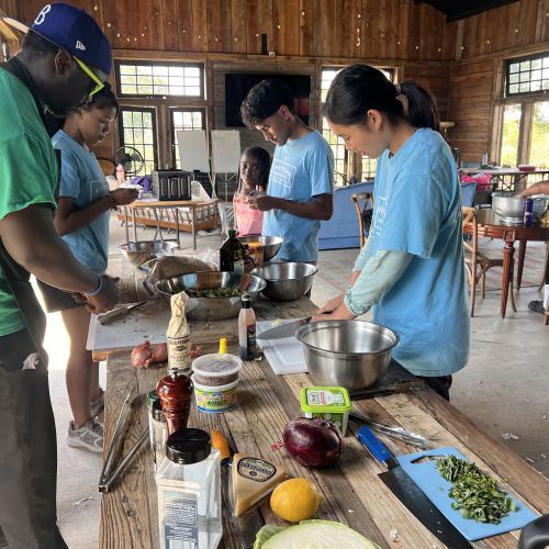 Chef Reggie Robinson, dressed in a green shirt, teaches students how to cook. On their work station are various cooking supplies and ingredients, some of which have already been prepared under Robinson’s careful instruction. In the background are wooden tables, where another student prepares food on their own.