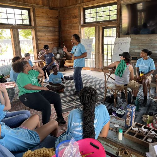 The Program Director, Juleon Lewis, stands in the middle of a cabin living room and instructs a group of students that form the Seed Crew and Assistant Crew Leaders. These students gather in seats around him and listen attentively as Lewis gestures throughout his speech. Bright sunlight and glimpses of green trees can be seen through the cabin windows.