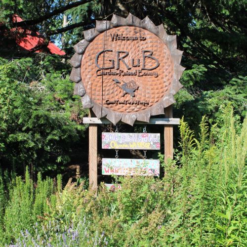 GRuB’s famous sign stands tall above thick, bright foliage. Its unique design reflects the shape of a sunflower, with GRuB’s logo placed proudly in the center. Behind the sign are rustling trees; a glimpse of a building’s orange rooftop can be seen between the leaves.