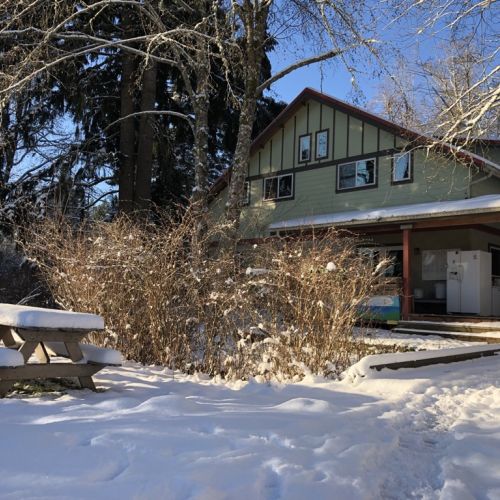 A snow-covered path opens up to reveal the GRuB Farmhouse, which is nestled in a circle of trees. There is a wooden picnic table with benches, also covered by a pristine layer of snow. Footprints can be seen on the snowy ground, reflective of GRuB’s many members who frequent the farmhouse throughout the day.