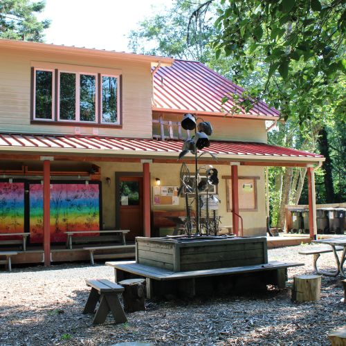 One of GRuB’s buildings, surrounded by green trees that shine brighter under the spring sunlight. The building’s windows are a colorful rainbow, which makes the environment feel more inviting. Patio tables and chairs are set up underneath the dappled shade of the trees overhead.