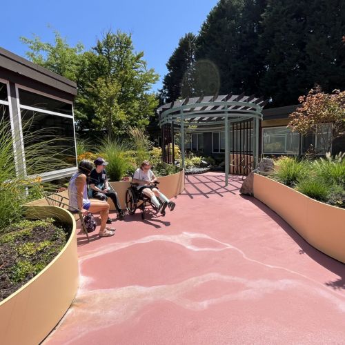 Three people—one of them in a wheelchair—are seated in a garden area that resembles an eddy, where fresh air is gathered; rows of planters border the walkway leading up to the seating area. The trio enjoys the spring bloom and the clear, sunny sky.