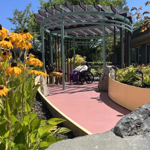 Another view of the pavilion, which is bordered by a stone wall in the foreground. This wall, combined with plantings, creates an area outside of the indoor hospice units for residents to enjoy. A woman in a wheelchair sits beneath the pavilion.