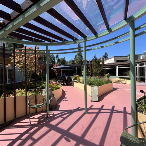  A view of the rest of the garden from the pavilion’s point of view. In the center is a raised planter bed, which is blooming with greenery. Residential buildings line either side of the garden walkway.