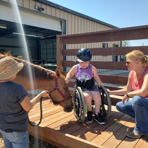 Two women support a child in a wheelchair on an accessible ramp while they pet the muzzle of a horse. The child is wearing a riding helmet. 