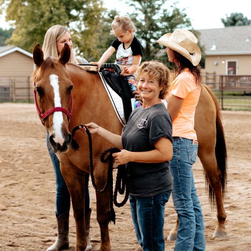 A young child is sitting on top of a horse with the support of three women. One woman is holding the reins, smiling warmly at the camera. The other two women stand beside the horse, helping the child in the saddle. The horse is brown with a white stripe down its muzzle. 