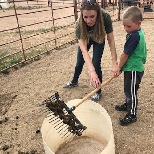 An adult woman and young boy in green shirts scooping animal manure into a white barrel with a pitchfork.