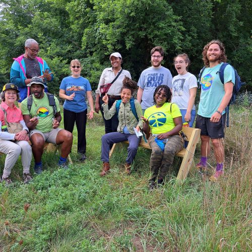 A group of participants gather closely to pose for a photo. Some people sit on wooden benches while others stand behind them. The scenery around them is full of green, as there are verdant trees behind them and tall wild grass surrounding them. The participants convey their admiration of the land through wide smiles.