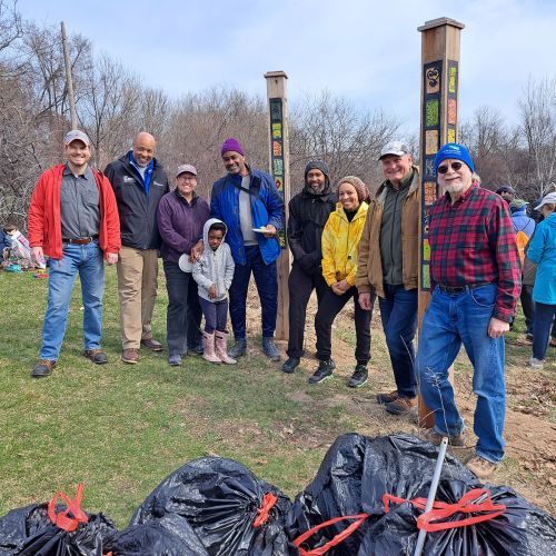 A group of participants dressed in warm layers pose for a picture in the middle of a park, which they had been working to clean up. Black trash bags are lined up as signs of the participants’ efforts to beautify their community. Behind the group are more people who were part of the cleanup, as well as barren trees in the far background.