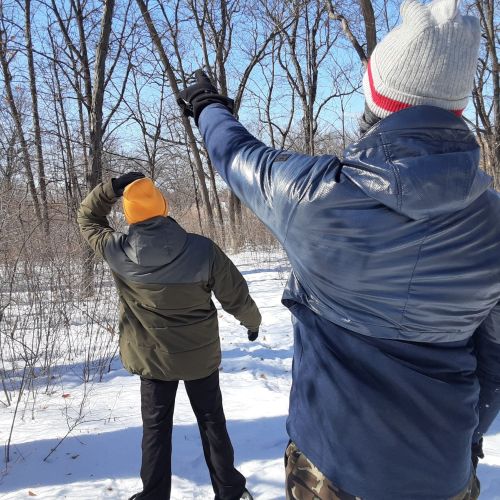 Two people in winter clothes enjoy a crisp, snowy day. The person in the foreground is pointing to something in the distance, which the second person is trying to find. Around them are thin, brittle trees that jut out from the snow-covered ground. The sky peeking through the trees is clear and light blue.