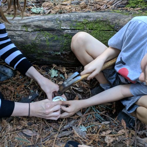 On a forest floor of leaves and twigs, next to a log, a child hammers a nail into a metal lid, helped by an adult who holds the nail with them. This experience builds attention, fine motor control, and a sense of pride and achievement when the project is completed.