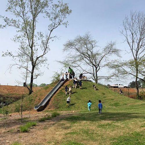 A crowd of children are climbing up and gathering to form a line behind a tall slide, which is propped up on a green hill.