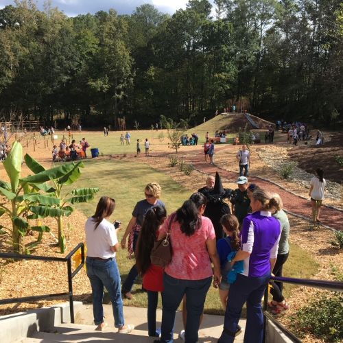 On grand opening day, a group of adults and children are looking at a view of an open area encircled by tall trees. Other people are scattered throughout the area and the earthen mound with slides are in the right background.