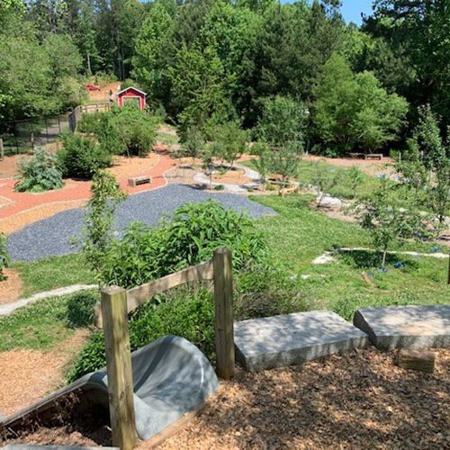 Standing atop the earthen mound near the entrance to a slide provides a clear view of the park that includes healthy trees, open grassland, and a play area.