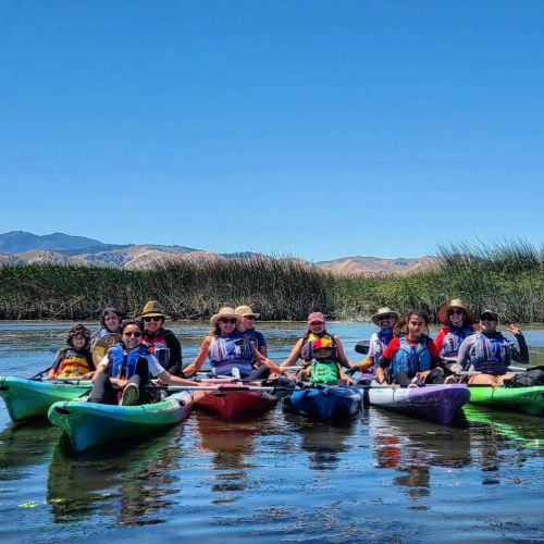 A group of people are kayaking down the CA Delta. The water itself is calm aside from the ripples caused by the kayaks. The blue sky above boasts no traces of clouds, promising a sunny day ahead for the participants.