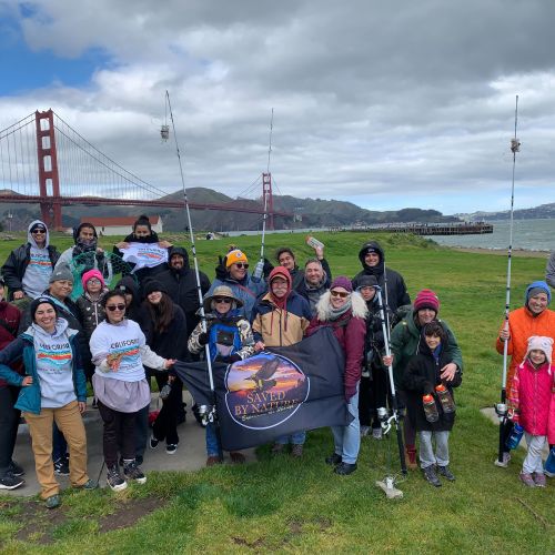 People are bundled up in warm clothes as they go crab fishing at Torpedo Pier during a cloudy day, with some holding up fishing rods. They all stand near the edge of a grassy river bank, with the Golden Gate Bridge looming tall in the background.