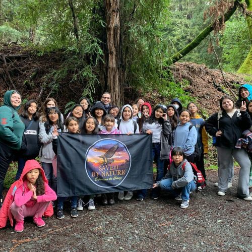 A large group of youths gather to take a picture for the Boys & Girls Club after-school hike. Behind the group are a wall of trees and foliage that altogether create the vibrant green backdrop of a rich, verdant forest.