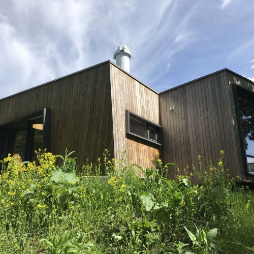 The exterior of the Outdoor Care Retreat. Its wooden structure stands tall against a summer blue sky; glass windows provide the cabin ample lighting. Photo credit: Maren Lindheim.