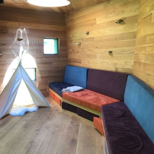 The interior family room, which boasts wooden flooring, walls, and ceiling. Couches, with blue, orange, and brown cushion are lined up against two walls for family members to sit, and a small blue hut with a white blanket inside the hut is set up for children to play in. Photo credit: Åshild Hauge