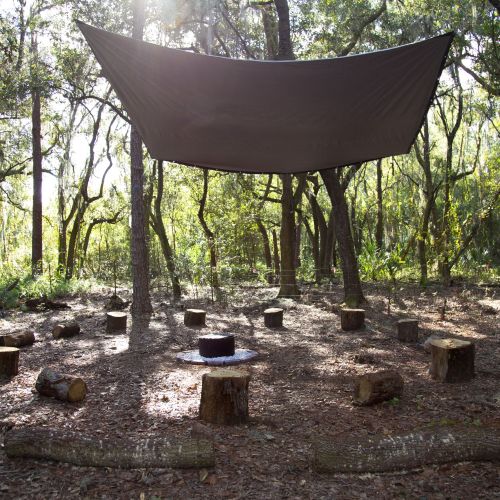 A clearing in a woodland is set up for community gathering, with 12 wooden stumps circling a space for a campfire. There is a canopy suspended above that provides some shade coverage and outside the circle, more logs are arranged for seating.